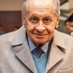 Marco Fidel Rocha en su oficina, rodeado de libros y diplomas, mirando hacia la ventana con una expresión reflexiva.