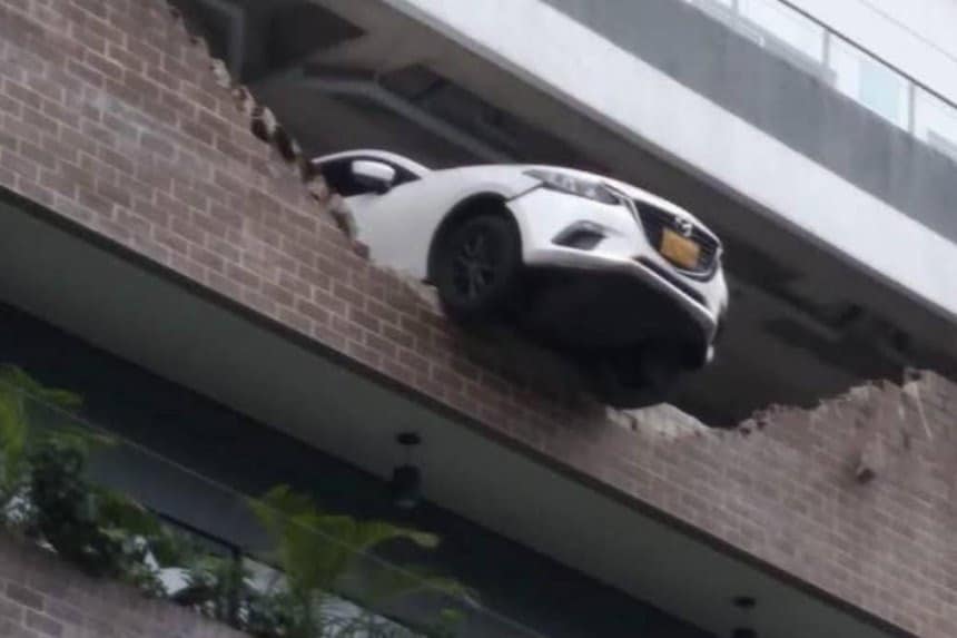 Vehículo rojo al borde de un parqueadero, con un muro dañado detrás y una mujer sorprendida en el interior.